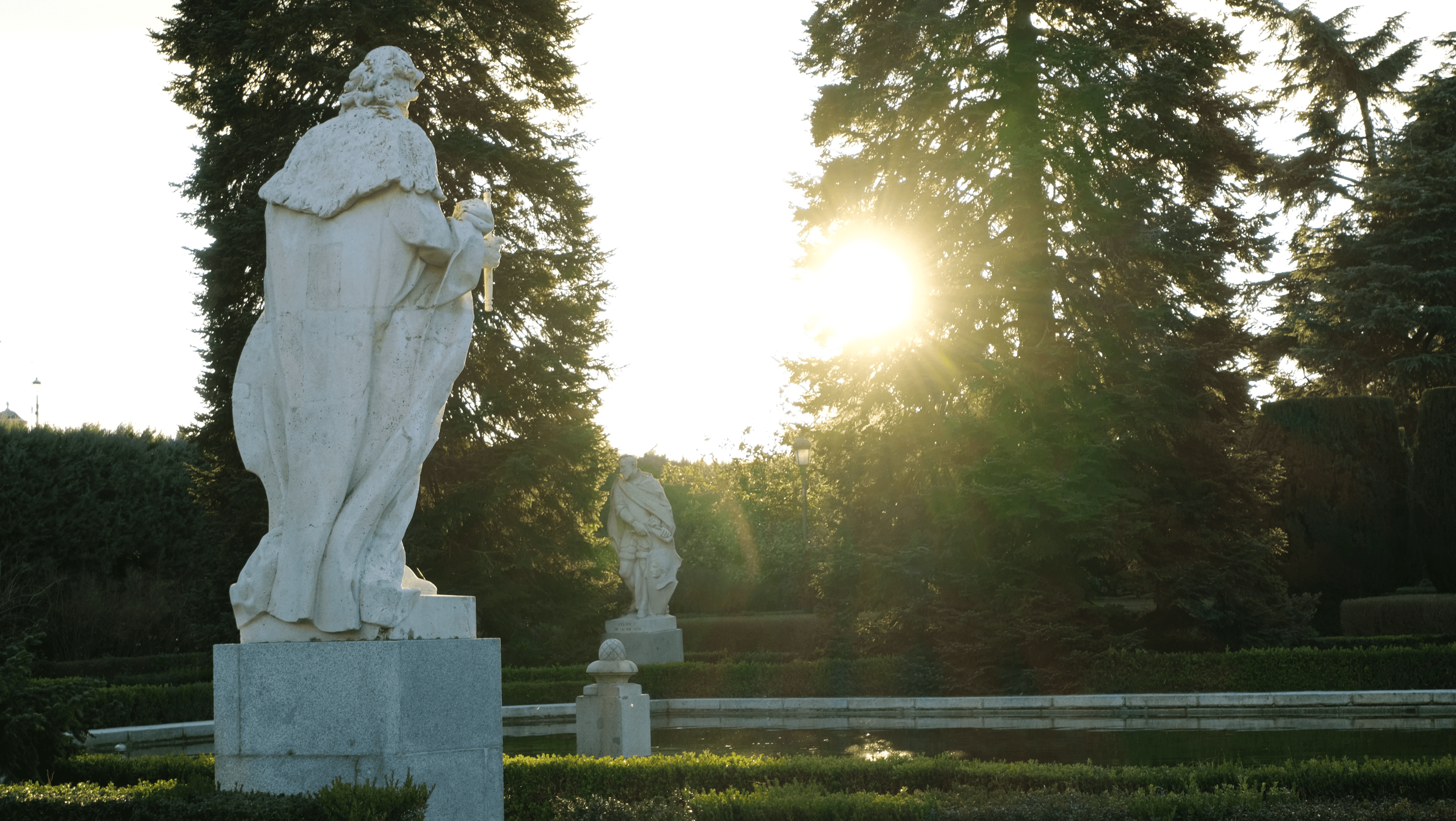 Stone statues in a formal garden, backlit by low sun through the trees
