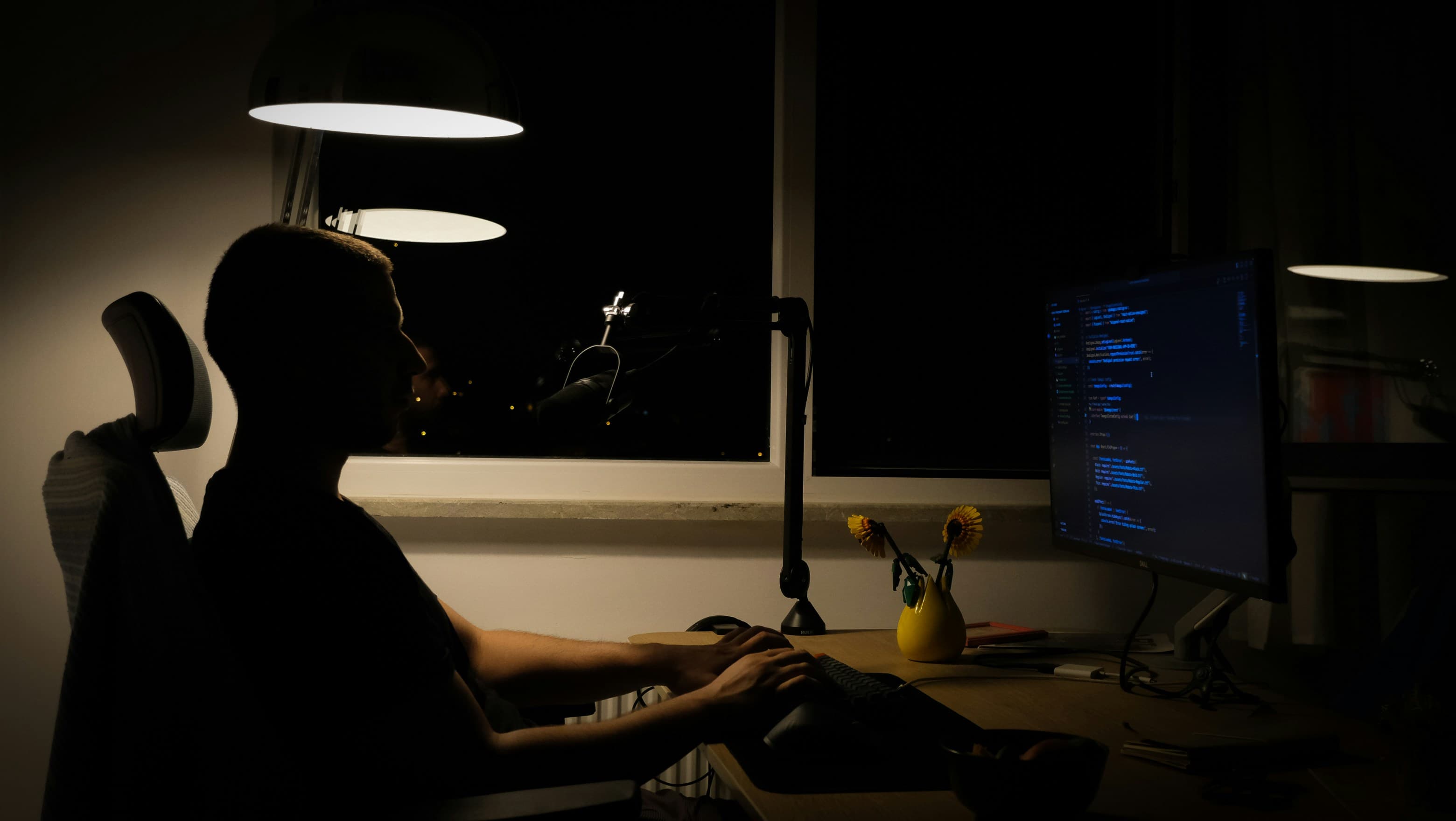 A person typing at the desk at night, lit by a single lamp, with code on the monitor