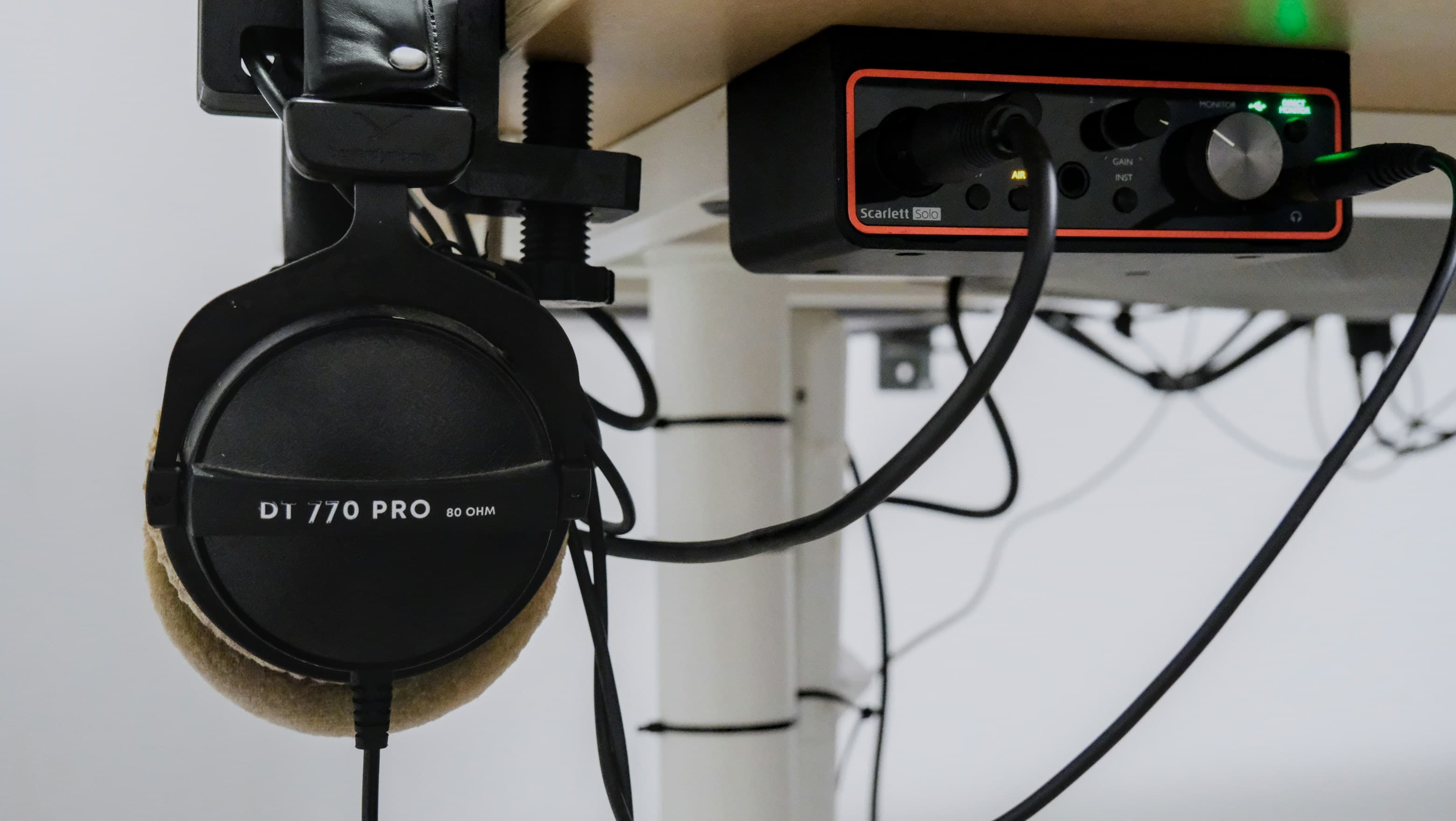 Closed-back studio headphones hanging from a hook beside an audio interface, both fixed under a desk