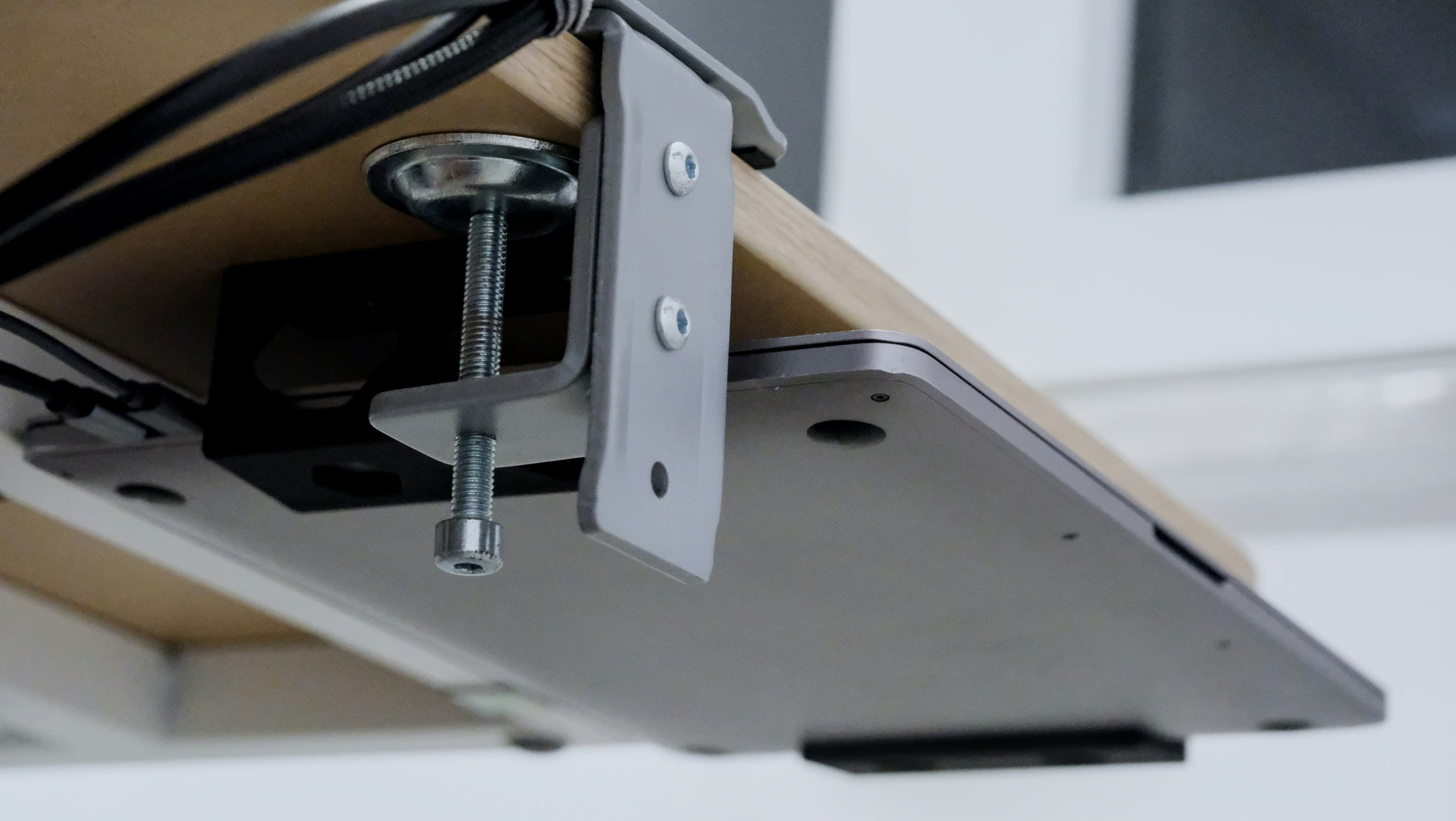 A metal clamp fixed under a wooden desk top, holding a closed laptop on a tray