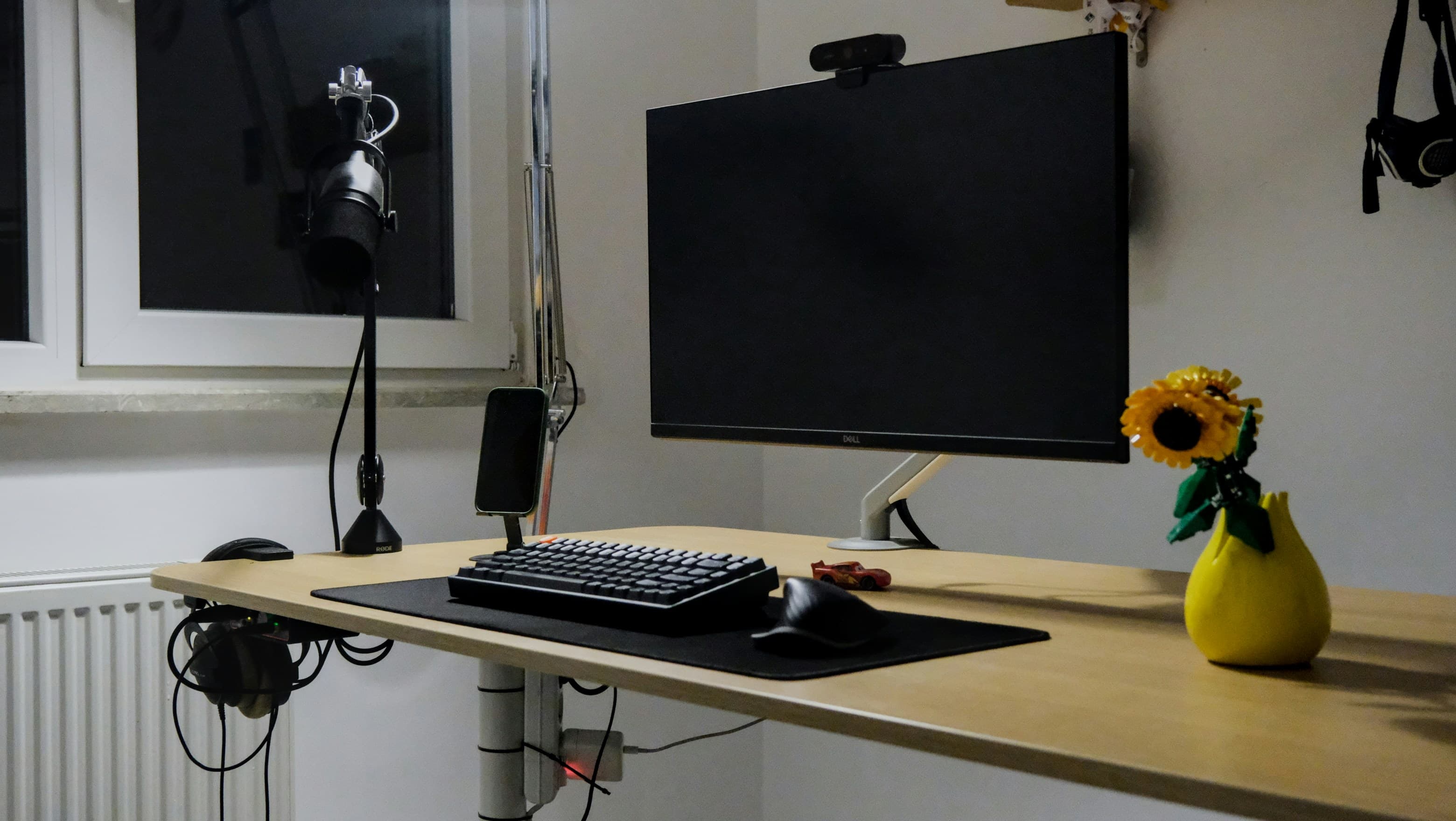 A desk with a monitor on an arm, a mechanical keyboard, a microphone on a boom and a yellow vase